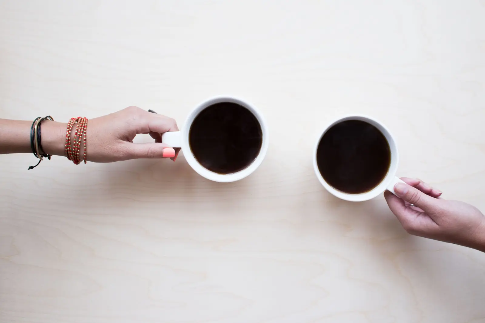 Two people's hands holding white mugs of coffee across a pale wooden table