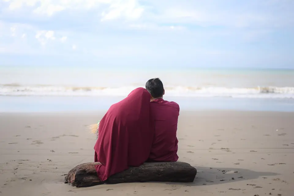 A couple wrapped in one shawl, sitting close on a beach and looking out at the sea