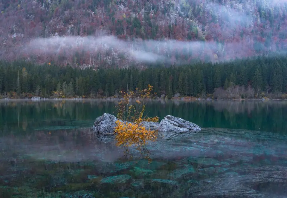 Clear, still water over a stony lake bed with mist on the forested hills beyond