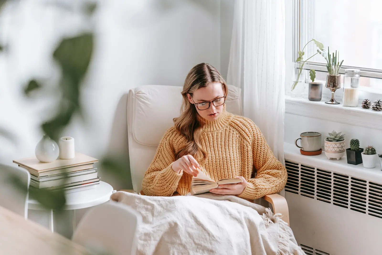 A young woman reading in a white armchair under a blanket beside a sunny window