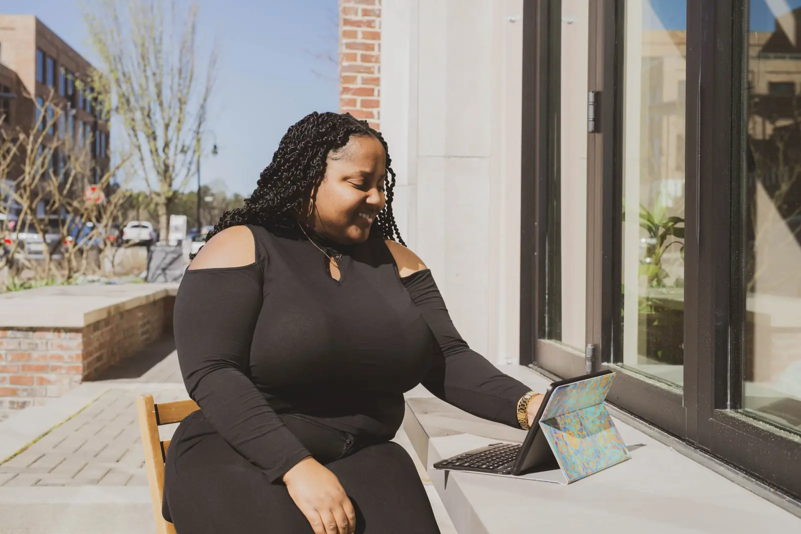 A woman sitting outside a sunlit building, smiling at the tablet on her lap